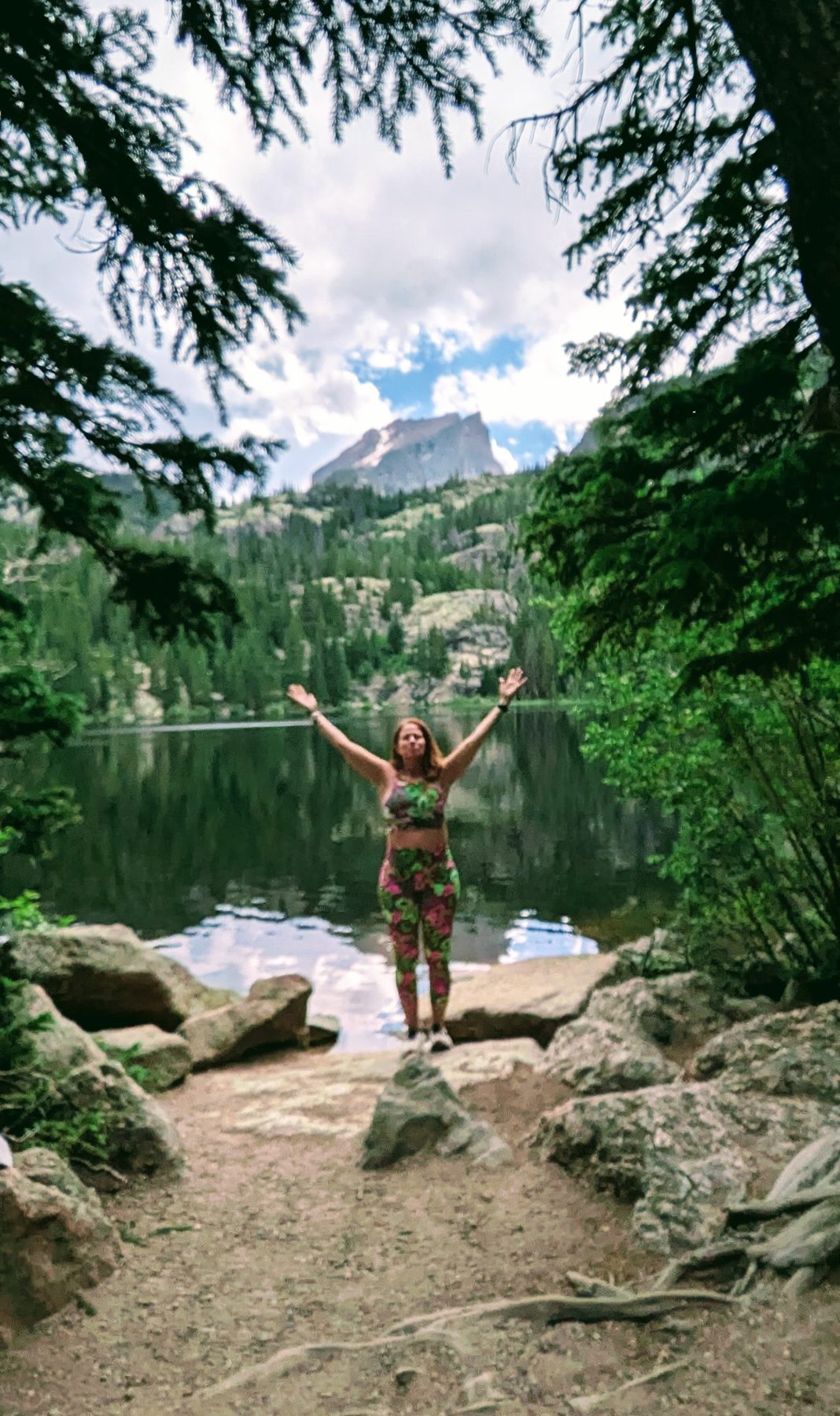 Marybeth with arms raised in celebration beside a mountain lake framed by pine trees